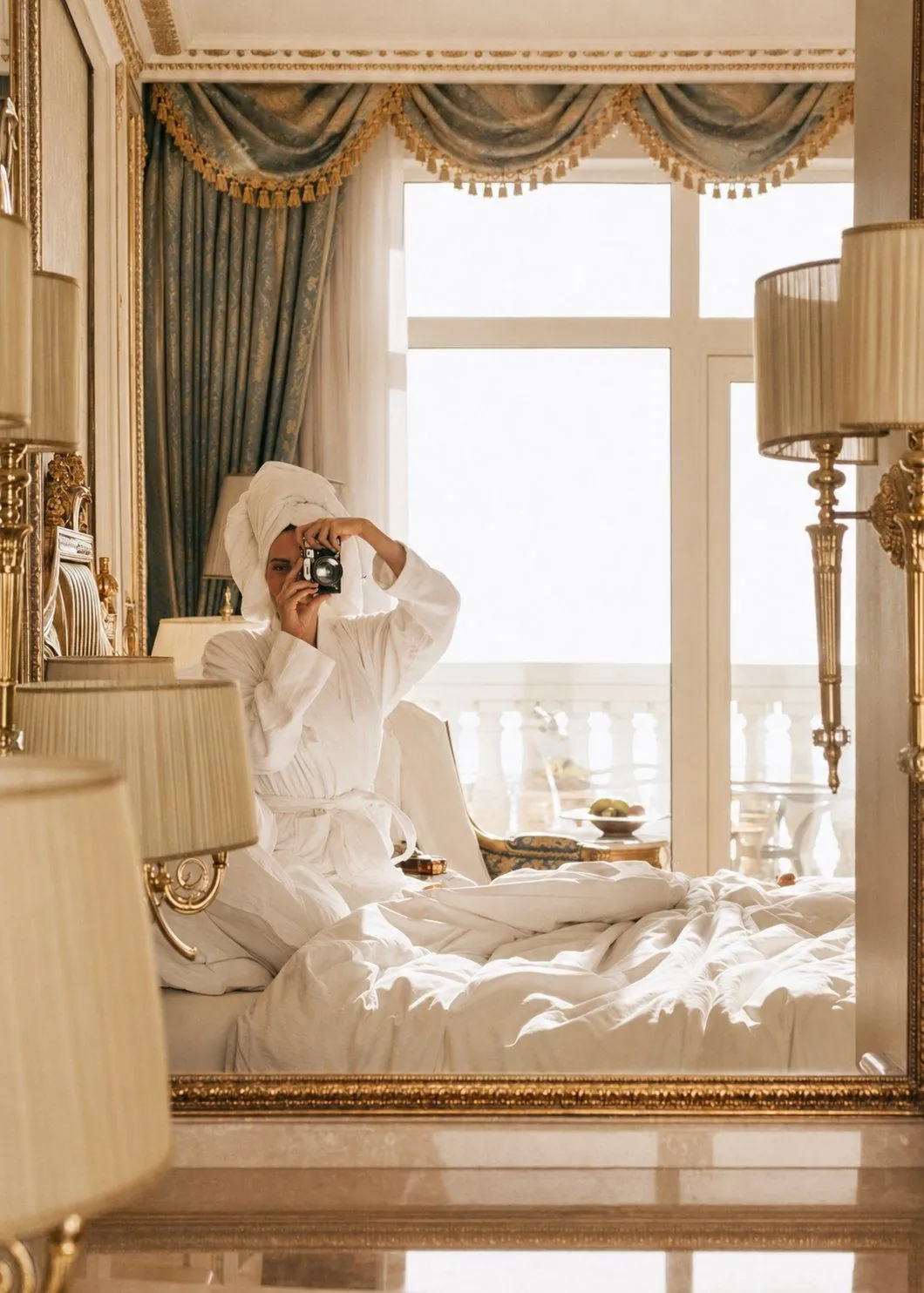 A guest in a bathrobe photographing herself in the mirror of a grand hotel room, morning light through the balcony doors