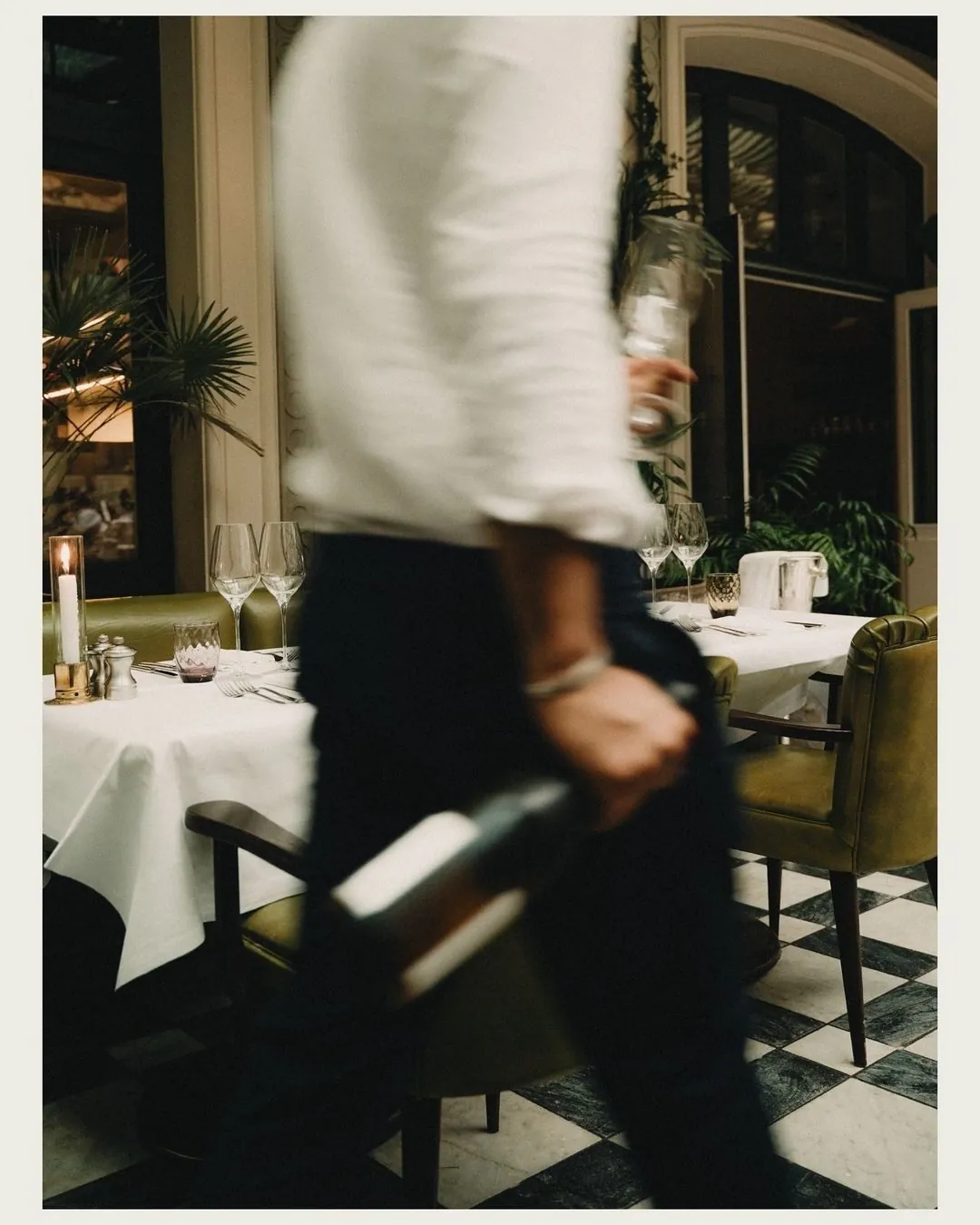 A waiter crossing a dining room mid-stride, tray in hand, the room slightly blurred around him