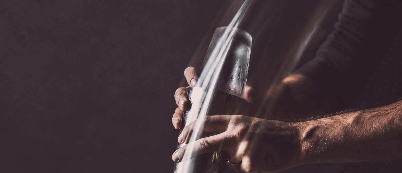 A pair of hands working a cocktail shaker at speed, the motion visible, the room dark behind