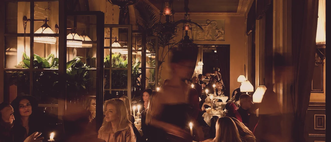 A candlelit dining room at capacity, one figure crossing the frame in motion blur
