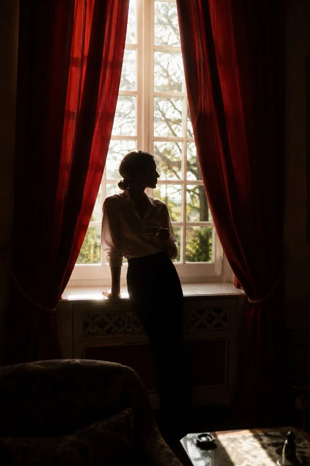 A woman silhouetted against a tall window between heavy red curtains, holding a cup